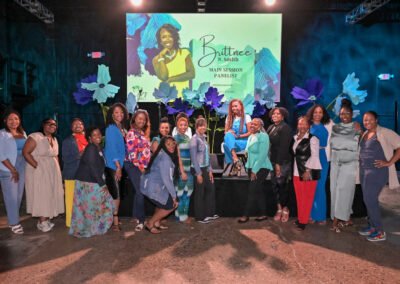 A group of women poses onstage at an event with a panelist sign behind, under bright lighting and floral decorations.