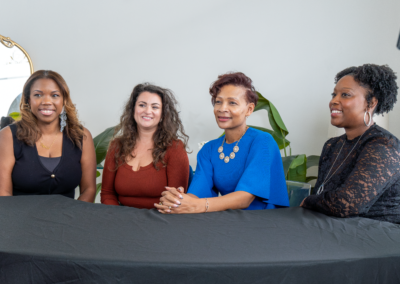Four women sitting at a table with a black tablecloth, smiling, with plants and a mirror in the background.