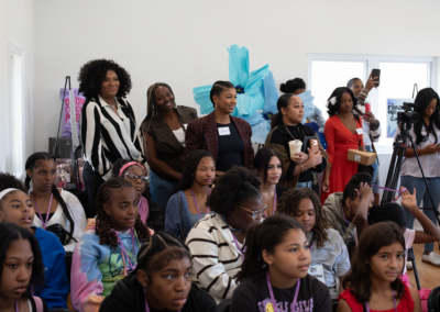 A diverse group of people, mostly young women, sitting and standing in a room, engaged in an event or workshop.