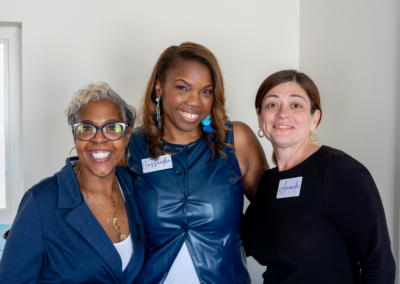 Three women smiling together indoors, wearing nametags and casual attire.