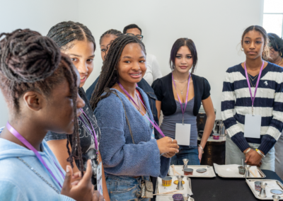 Group of young people with lanyards gathered around a table with trays and tools in a bright room.
