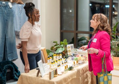 Two women converse at a market stall displaying skincare products, with jeans and plant decor in the background.