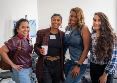 Four women smiling at a gathering, wearing name tags, with one holding a cup, standing against a white wall.