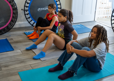 Three kids sit on yoga mats in a room, facing forward, with trampolines and a closed door in the background.