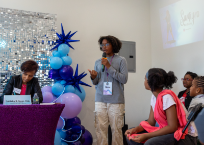 A person speaks into a microphone at a table, with attendees listening, in front of blue and purple balloon decorations.