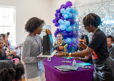 Person receives a gift at an event with an audience and vibrant balloon decorations in the background.