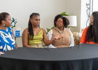 Four women sitting at a table, engaged in a discussion, with plants and decor in the background.