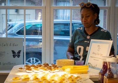 Chef in a sports jersey prepares food at a cafe event, with a sign for "Bites by Chef Zigga" and plates of appetizers.