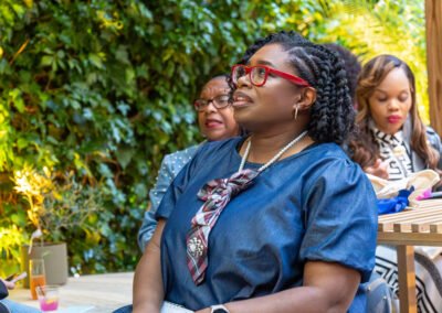 Woman with red glasses and pearl necklace at an outdoor gathering, surrounded by greenery and two other women.