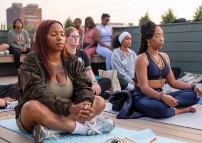 Group of people meditating on a rooftop, sitting cross-legged on yoga mats, with a cityscape background.