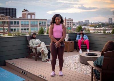 Woman in pink top stands on a rooftop terrace with three seated individuals, evening cityscape in the background.