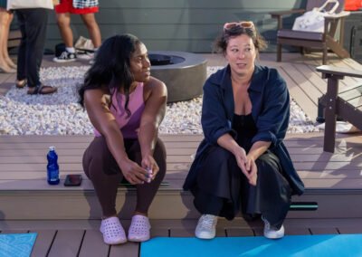 Two women sitting on a deck, chatting, with yoga mats and a water bottle beside them. Others stand in the background.