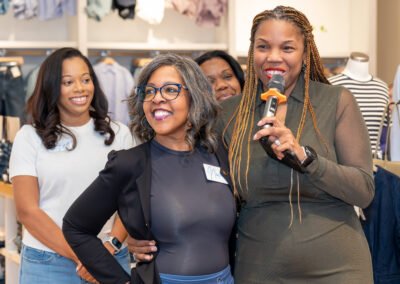 Women smiling at a gathering in a clothing store; one holds a microphone.