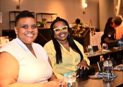 Two women smiling at a table in a warmly lit room, with glasses and notepads in front of them.