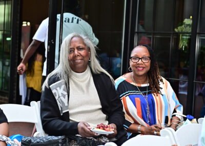 Two women seated outdoors, one holding a plate of grapes, both smiling, with a glass door in the background.