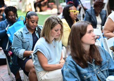 A group of people seated outdoors, focused on an event, with blue bags visible in the background.