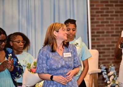 A woman in a blue dress with a name tag stands smiling among others, one holding flowers, in a decorated event space.