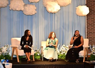 Three women seated onstage holding microphones, with cloud decorations and floral arrangements in the background.