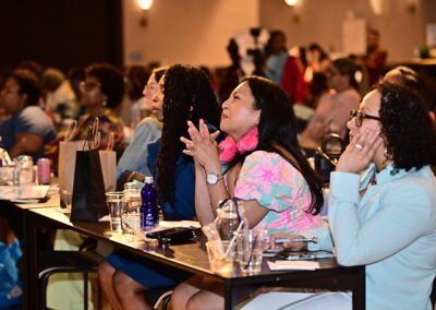 Audience attentively listens during a conference, seated at tables with drinks and notepads, in a warmly lit room.