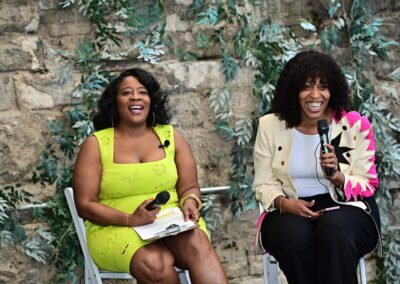 Two women laughing and holding microphones, seated against a leafy stone wall backdrop.