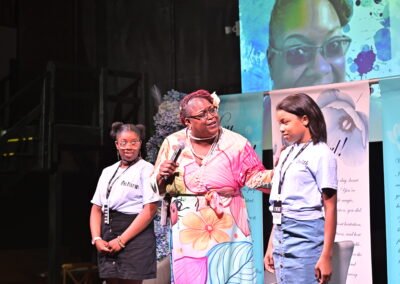 Three people on a stage. A person with a microphone speaks to two others wearing "event staff" tags, against a floral backdrop.