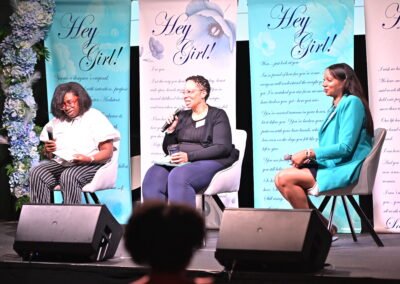 Three women seated on stage with microphones at a "Hey Girl!" event, floral backdrop, and printed banners.