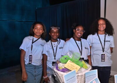 Four smiling young girls in matching event t-shirts pose together indoors beside a basket filled with various items.