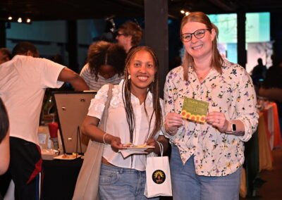Two women smiling at an indoor event, holding a thank you card and gift bag, with people mingling in the background.