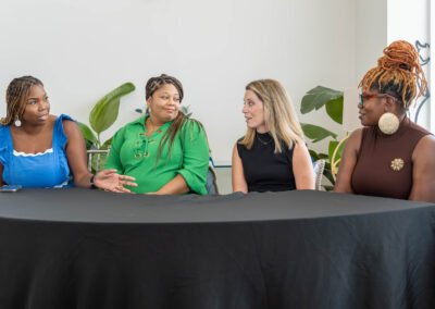 Four women engaged in discussion around a black-covered table, with green plants in the background.