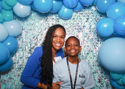 Two people smiling in front of a blue balloon arch and sparkly backdrop at an event.