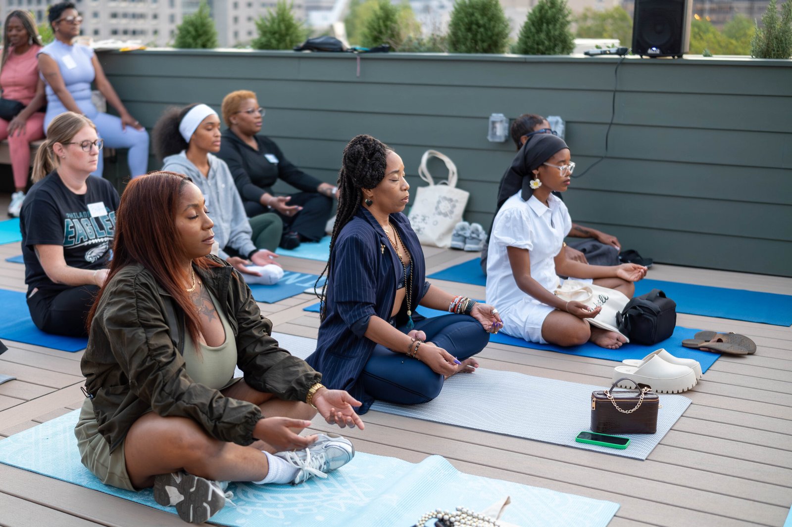 A diverse group of people meditate on yoga mats at an outdoor rooftop space, focusing calmly with eyes closed.