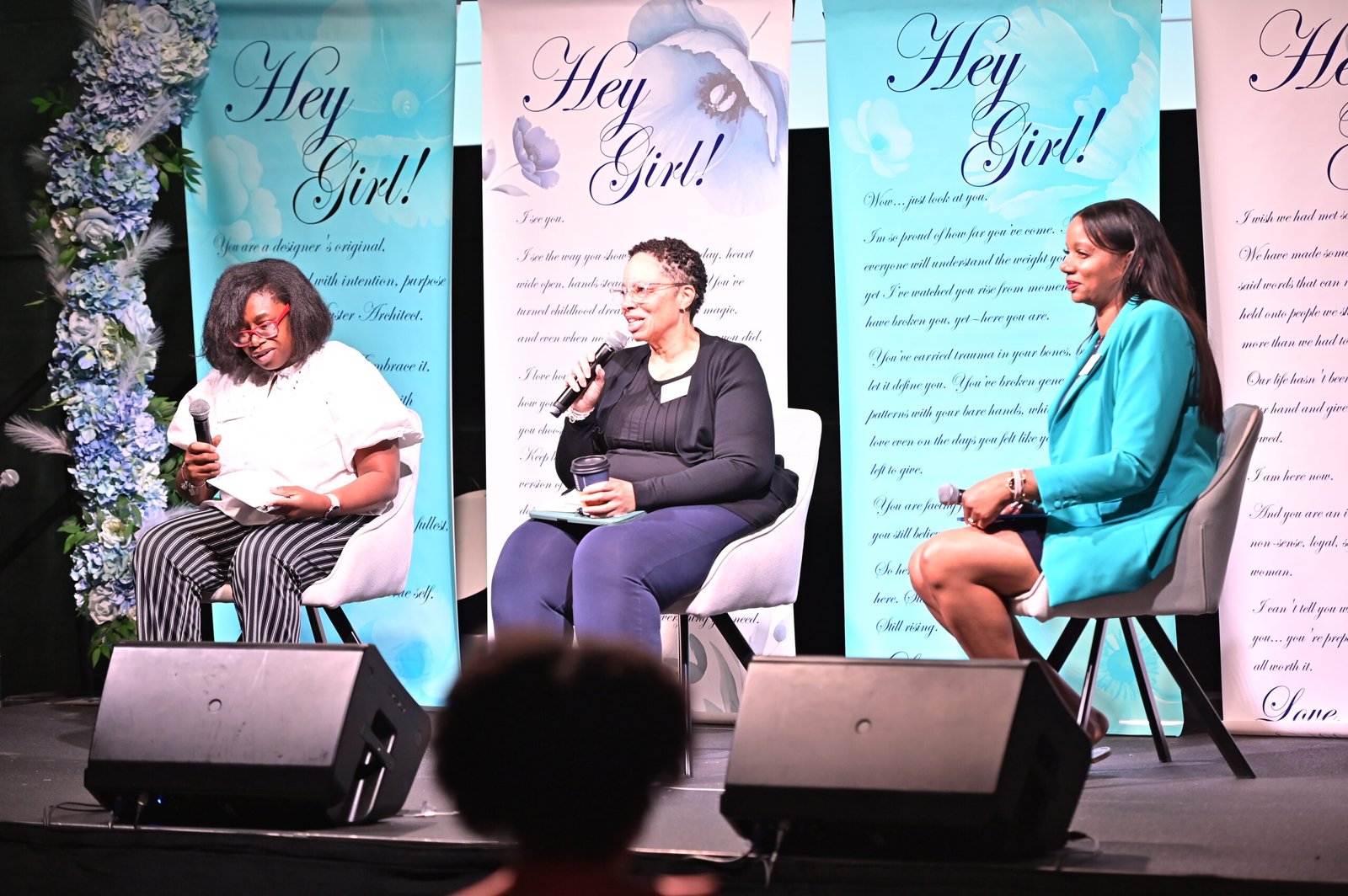 Three women sit in chairs holding microphones, engaging in a panel discussion against "Hey Girl!" banners.