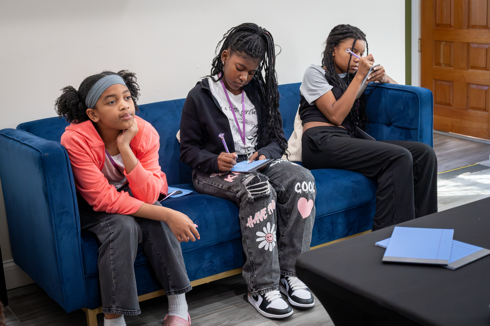 Three girls sitting on a blue couch, writing in notebooks with pens.