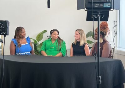 Four women sitting at a round table having a discussion, surrounded by studio lighting equipment and plants.